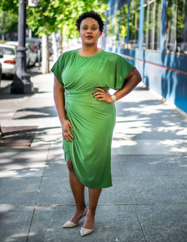 Full length portrait of Niema Jordan standing hand on her hip on an Oakland California sidewalk, dappled light, buildings, trees. Niema wears a gold watch, an asymmetrically flowing green dress to her knees, delicate hoop earrings, sharp flats. Her nails are painted and her hair is like a crown. 