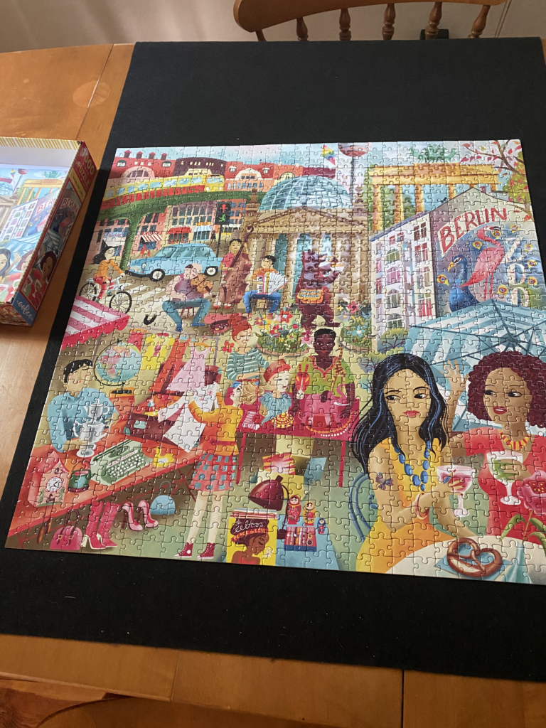 A completed puzzle of an illustrated Berlin flea market scene on a cardboard mat on a table. 
