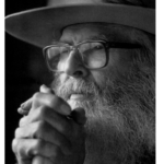 Black and white portrait of William Oliver Everson close up with hands clasped in front of his face wearing a long beard, dark rimmed glasses, and a wide brimmed hat.