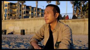 Tony sits on the beach at golden hour looking at the ocean. Palm trees and concrete in the background.