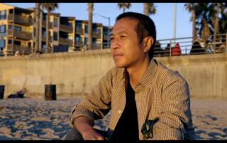 Tony sits on the beach at golden hour looking at the ocean. Palm trees and concrete in the background.